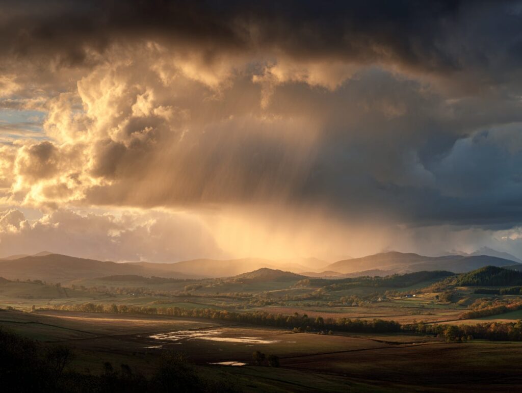 A dramatic landscape photograph of golden sunlight breaking through dark storm clouds over a misty valley of rolling hills, distant mountains, and fields below — symbolizing clarity and steadiness returning to a turbulent inner sky, a bridge to the article's central claim that a regulated nervous system is what lets a leader think clearly through pressure and uncertainty.